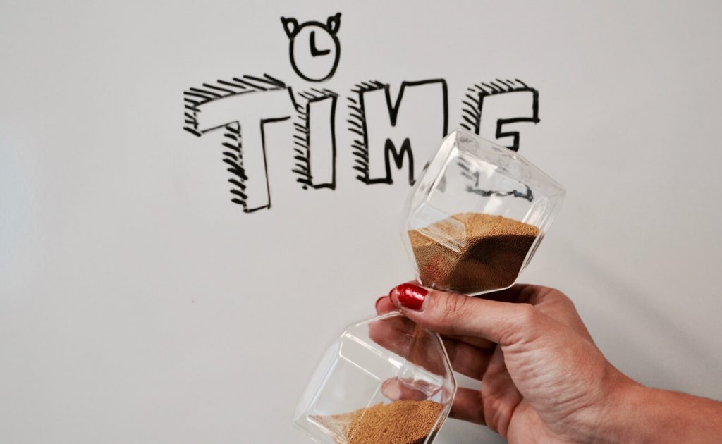a whiteboard with "time" written on it behind a hand holding an hourglass